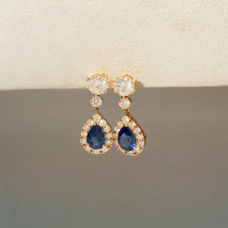 Front view of sapphire and diamond drop earrings in yellow gold, featuring pear-shaped blue gemstones framed by sparkling diamonds. Set against a neutral backdrop, the design exudes timeless elegance.