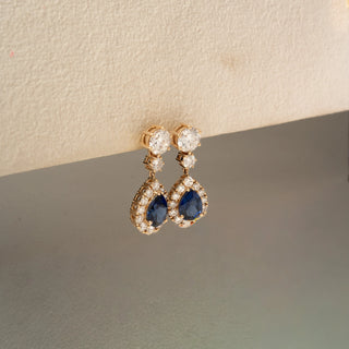 Side view of sapphire and diamond drop earrings in yellow gold, featuring pear-shaped blue gemstones framed by sparkling diamonds. Displayed against a neutral backdrop with soft lighting.