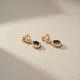 Angled view of sapphire and diamond drop earrings in yellow gold, featuring pear-shaped blue gemstones framed by sparkling diamonds. Set against a beige backdrop, the design radiates refined elegance.