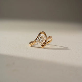 Front view of a yellow gold ring featuring an oval-cut solitaire diamond framed by an elegant crossover setting, creating a modern and sculptural design against a soft neutral background.