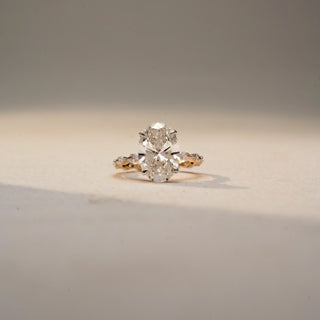 Front view of an oval diamond engagement ring in 14 KT Yellow Gold, featuring an oval-cut lab-grown diamond with marquise and round diamond accents along the band, displayed against a beige backdrop.