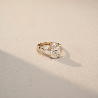 Angle view of an oval diamond engagement ring in 14 KT Yellow Gold, featuring an oval-cut lab-grown diamond with marquise and round diamond accents along the band, displayed against a beige backdrop.