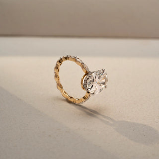 Side view of an oval diamond engagement ring in 14 KT Yellow Gold, featuring an oval-cut lab-grown diamond with marquise and round diamond accents along the band, displayed against a beige backdrop.