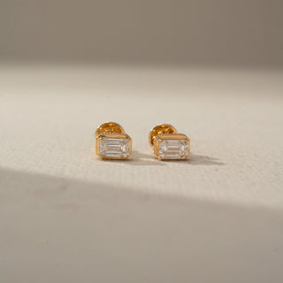 Front view of a pair of gold stud earrings featuring emerald-cut clear gemstones in sleek bezel-style settings, displayed on a neutral surface with soft natural light.