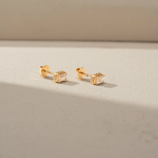 Side view of a pair of gold stud earrings featuring emerald-cut clear gemstones in raised settings, displayed on a neutral surface with soft natural light and subtle shadows.