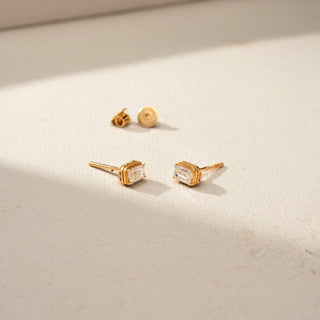 Angled view of a pair of gold stud earrings featuring emerald-cut clear gemstones in ornate basket settings, displayed with screw-back closures on a softly lit neutral surface.