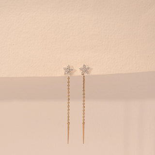 Front view of a 14KT yellow gold earrings featuring floral diamond details with delicate chain drops, set against a beige background.
