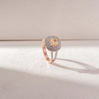 Angled view of Cushion Halo Diamond Ring in 14KT Rose Gold, featuring a cushion-cut yellow diamond with double pavé halo and split diamond band. Set on a soft beige surface, it radiates brilliance and depth.
