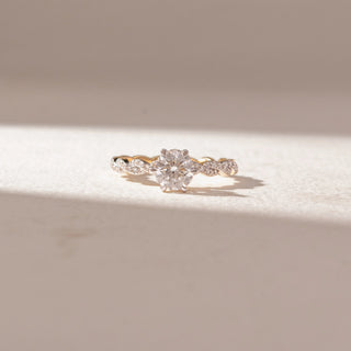 Front view of round diamond solitaire ring in yellow gold with a twisted pavé band, set on a soft neutral surface, highlighting elegant curves and brilliant, light-catching sparkle.
