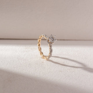 Angled view of round diamond solitaire ring in yellow gold with a twisted pavé band, set on a soft neutral background, highlighting braided design and radiant, light-catching sparkle.
