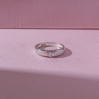 Top view of a white gold diamond ring, featuring a centred round-cut diamond between smooth polished bands. Captured on a soft pink backdrop, highlighting clean elegance and subtle sparkle.
