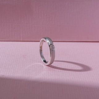 Angled view of a white gold diamond ring, featuring a centred round-cut diamond set within sleek polished bands. Captured on a soft pink backdrop, highlighting refined minimalism and shine.
