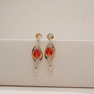 Front view of yellow gold drop earrings featuring pear-shaped red ruby centre stones framed by curved diamond accents with pear diamond drops, displayed on a soft neutral background.