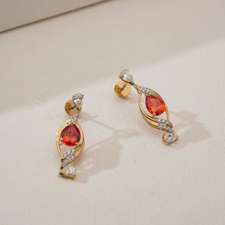 Angled view of yellow gold drop earrings with pear-shaped red ruby centre stones, curved diamond accents and pear diamond tips, placed on a soft neutral surface.