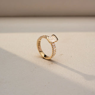 Angled view of an emerald-cut solitaire ring in 14 KT Yellow Gold, featuring an emerald-cut lab-grown diamond in a raised setting with graduated baguette diamonds along the band, displayed against a beige backdrop.