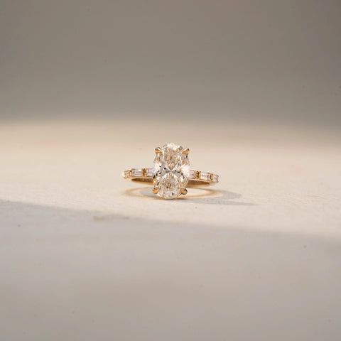 Front view of an oval three-stone ring in 14 KT Yellow Gold, featuring a central oval-cut lab-grown diamond flanked by tapered baguette diamonds, displayed against a beige backdrop.