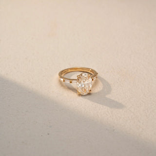 Side view of an oval three-stone ring in 14 KT Yellow Gold, featuring a central oval-cut lab-grown diamond flanked by tapered baguette diamonds, displayed against a beige backdrop.