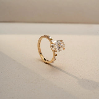 Angled view of an oval three-stone ring in 14 KT Yellow Gold, featuring a central oval-cut lab-grown diamond flanked by tapered baguette diamonds, displayed against a beige backdrop.