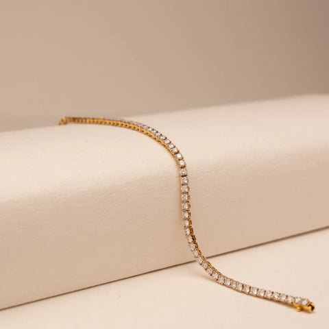 Angled view of Reia’s 14 KT Yellow Gold Timeless Elegance Diamond Tennis Bracelet. Captured on a cream backdrop, it highlights the continuous, shimmering line of round-cut diamonds.