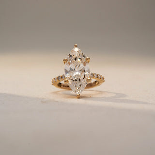 Front view of a marquise diamond engagement ring in 14 KT Yellow Gold, featuring a marquise-cut lab-grown diamond with pavé-set diamonds along the band, displayed against a beige backdrop.
