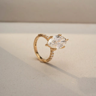 Angled view of a marquise diamond engagement ring in 14 KT Yellow Gold, featuring a marquise-cut lab-grown diamond with pavé-set diamonds along the band, displayed against a beige backdrop.
