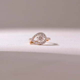 Front view of a diamond halo ring in yellow gold, featuring a round center stone framed by a pavé swirl design. Captured against a soft beige backdrop, the design reflects elegance and radiant sparkle.
