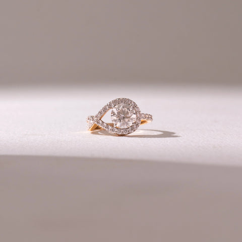 Front view of a diamond halo ring in yellow gold, featuring a round center stone framed by a pavé swirl design. Captured against a soft beige backdrop, the design reflects elegance and radiant sparkle.
