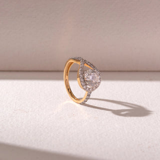 Angled view of a yellow gold diamond halo ring, featuring a round center stone framed by a pavé swirl design. Captured against a soft beige backdrop, the design reflects graceful sparkle and elegance.
