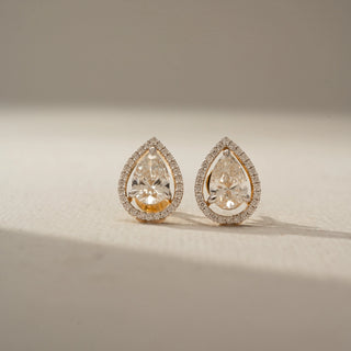 Front view of pear-shaped diamond stud earrings with a delicate halo of pavé diamonds, set in yellow gold and displayed on a softly lit neutral surface.