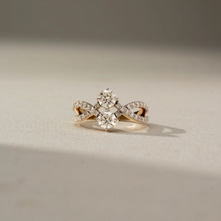 Front view of a diamond crossover ring featuring two round brilliant-cut diamonds and pavé-set diamond infinity-style bands, displayed on a soft neutral surface.