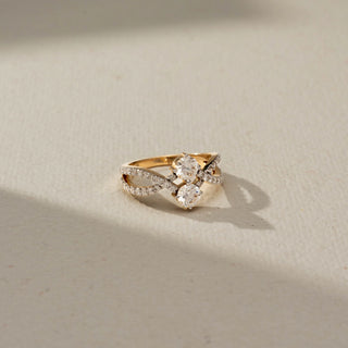 Top view of a diamond crossover ring featuring two round brilliant-cut diamonds and pavé-set infinity-style bands in yellow gold, displayed on a soft ivory surface with natural light.