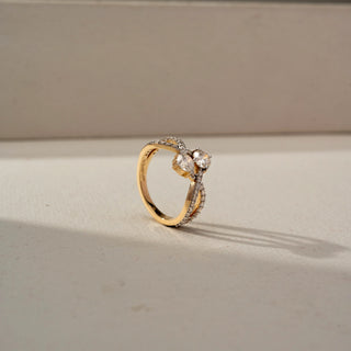 Angled view of a yellow gold diamond ring featuring a twin-stone floral-inspired centerpiece and a split pavé-set band, elegantly displayed on a neutral surface with soft natural light.