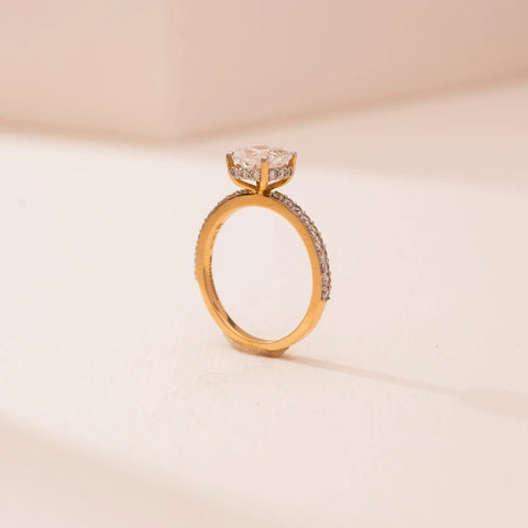 Side view of Reia’s 18 KT Yellow Gold Softglow Cushion Solitaire Diamond Ring, showing a round-cut stone on a pavé band. Set against pink, it highlights warm, symmetrical brilliance.