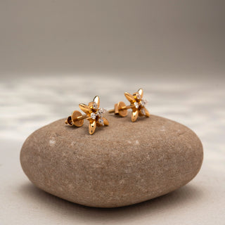 Side view of floral diamond stud earrings in 14 KT Yellow Gold, featuring polished petal motifs with a sparkling round diamond flower cluster at the center, displayed on a textured stone surface.