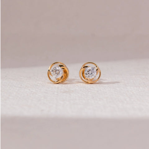 Front view of swirl diamond stud earrings in yellow gold, featuring centred round lab-grown diamonds encased in interlocking circular loops. Placed on a soft beige surface, the design reflects modern elegance.
