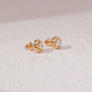 Angled view of swirl diamond stud earrings in yellow gold, featuring centred round lab-grown diamonds encased in interlocking circular loops. Placed on a textured beige surface, the design casts a soft shadow.

