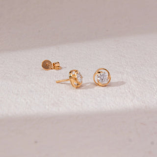 Top view of swirl diamond stud earrings in yellow gold, featuring centred round lab-grown diamonds encased in smooth circular loops. Placed on a textured beige surface, the design casts a soft shadow.
