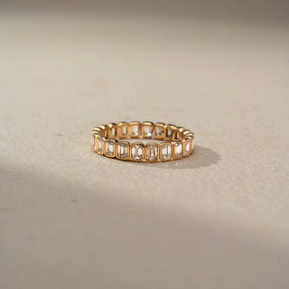 Front view of an emerald-cut diamond eternity band in 14 KT Yellow Gold, featuring bezel-set emerald-cut lab-grown diamonds encircling the entire band in a timeless design, displayed against a beige backdrop.
