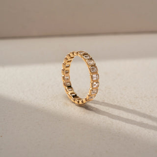 Angled view of an emerald-cut diamond eternity band in 14 KT Yellow Gold, featuring bezel-set emerald-cut lab-grown diamonds encircling the entire band in a timeless design, displayed against a beige backdrop.