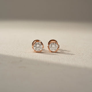 Front view of round diamond stud earrings in rose gold, featuring a modern four-petal floral-inspired setting that frames each sparkling center stone on a neutral backdrop.