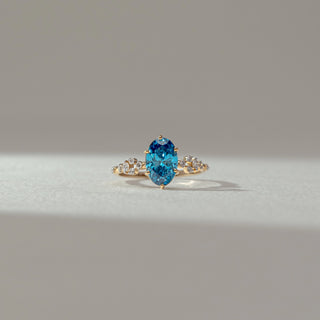 Front view of blue gemstone ring in yellow gold, featuring an oval-cut vibrant blue centre stone with round lab-grown diamond accents on the band. Set on a soft neutral backdrop, it reflects bold colour and elegance.