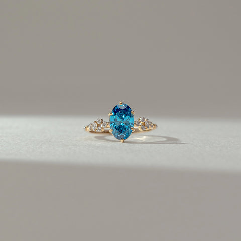 Front view of blue gemstone ring in yellow gold, featuring an oval-cut vibrant blue centre stone with round lab-grown diamond accents on the band. Set on a soft neutral backdrop, it reflects bold colour and elegance.