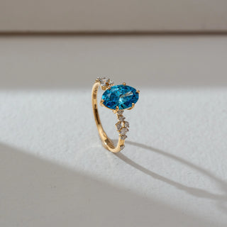 Side angled view of blue gemstone ring in yellow gold, featuring an oval-cut vibrant blue centre stone with round lab-grown diamond accents along the band. Set on a soft neutral backdrop, it highlights brilliance and colour.