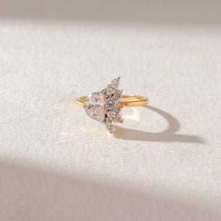 Top view of pear-shaped diamond cluster ring in yellow gold, featuring a central pear diamond with marquise and round accents, set on a soft neutral surface, highlighting a floral-inspired design and bright sparkle.
