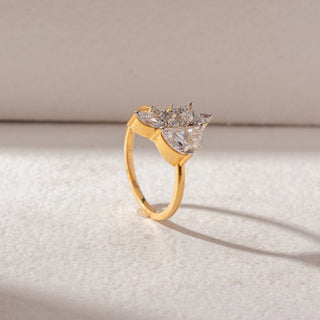 Angled view of pear-shaped diamond cluster ring in yellow gold, featuring a central pear diamond with marquise and round accents, set on a soft neutral surface, highlighting elevated setting and radiant sparkle.
