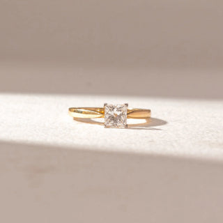 Front view of princess-cut diamond solitaire ring in yellow gold with a slim band, set on a soft neutral surface, highlighting sharp symmetry and clean, radiant brilliance.
