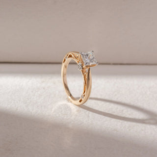 Angled view of princess-cut diamond solitaire ring in yellow gold with an intricate openwork band, set on a soft neutral background, highlighting detailed craftsmanship and radiant sparkle.
