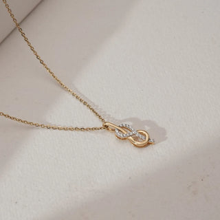 Angled view of Infinity Loop Golden Diamond Pendant in yellow gold, showcasing intertwined infinity curves lined with round lab-grown diamonds and a delicate dangling accent, placed on a textured beige surface.
