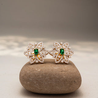 Front view of floral diamond stud earrings in 14 KT Yellow Gold, featuring pavé-set looping petals with emerald-cut green center stones, displayed on a textured stone surface.
