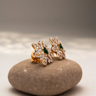 Side view of floral diamond stud earrings in 14 KT Yellow Gold, featuring pavé-set looping petals with emerald-cut green center stones, displayed on a textured stone surface.